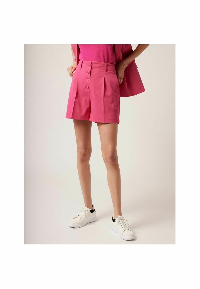 Shorts taille haute roses avec fermeture à boutons sur le devant, plis et finition texturée. Associés à des baskets blanches avec des accents noirs.