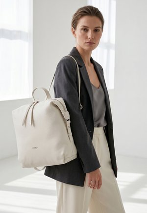 Donna che indossa un blazer scuro e pantaloni chiari con uno zaino in pelle grande color avorio chiaro in uno spazio interno luminoso e minimalista.