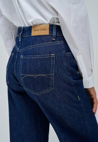 Jeans in denim blu scuro con un dettaglio di tasca posteriore cucita e un'etichetta in pelle beige con la scritta "SALSA JEANS" sulla cintura.