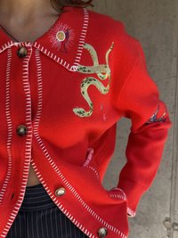 Piros kötött kardigán kontrasztos fehér varrással. Bonyolult zöld kígyó és nap hímzéssel rendelkezik, valamint barna gombos záródással.