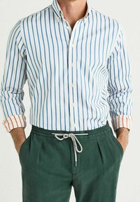 Camisa abotonada con rayas verticales azules y blancas, con acentos naranjas contrastantes en los puños; combinada con pantalones verde bosque con cordón.