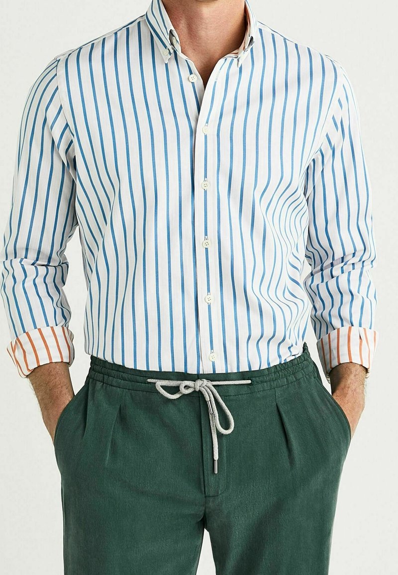 Camisa abotonada con rayas verticales azules y blancas, con acentos naranjas contrastantes en los puños; combinada con pantalones verde bosque con cordón.