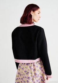 Jachetă neagră scurtă cu guler și tiv roz, care prezintă un material texturat, purtată peste o fustă cu model floral.