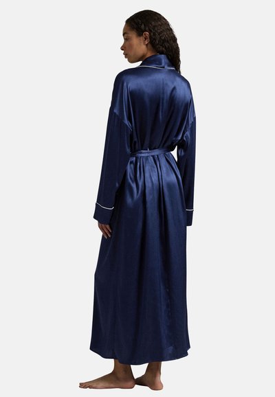 Vrouw staat blootsvoets, draagt een lange, marineblauwe satijnen badjas met een ceintuur, licht naar de zijkant gedraaid waardoor de achterkant en het profiel zichtbaar zijn.