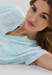Top de manga corta azul claro con cuello, que presenta un patrón de hojas en blanco y un bolsillo en el lado izquierdo. Textura de tejido suave.