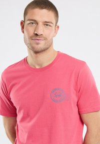 T-shirt en coton rose avec un col rond et un logo imprimé bleu sur la partie gauche de la poitrine. Manches courtes et coupe décontractée.