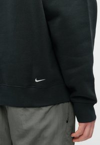 Sweatshirt preto com mangas longas, punhos canelados e um pequeno logótipo branco da Nike na bainha. O tecido parece suave e texturizado.