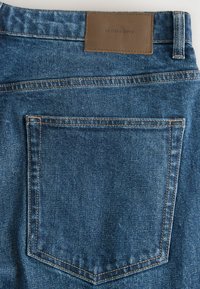 Sinise denimpükste tagatasku, millel on nähtavad õmblused ja pruun nahast silt, millel on kiri "DENIM SUPPLY" vöökoha peal.