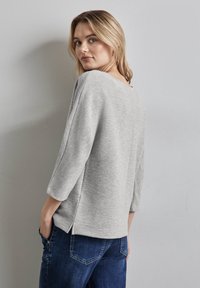 Top grigio testurizzato con vestibilità rilassata, scollo a V e maniche a tre quarti, abbinato a jeans blu scuro con fessure laterali.