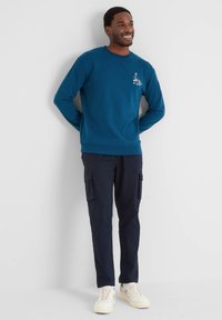 Smiling man in blue sweatshirt with graphic on chest, dark cargo pants, and white sneakers standing against a white wall with hands behind back.