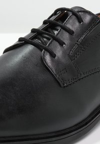 Chaussure en cuir noir avec une finition lisse, présentant des textures variées, un bout arrondi et un design classique à lacets avec de subtils détails de broderie.