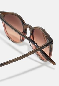 Ochelari de soare din fucsie cu lentile maronii degradate, formă clasică de ochi de pisică, brațe subțiri și marcajul Ray-Ban pe un templu.