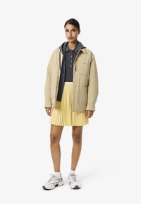 Beige vadderad jacka över en mörkgrå polo, parat med en plisserad gul kjol och vita sneakers. Slät textur, avslappnad design.