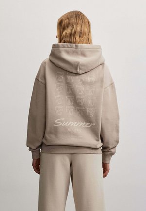 Person med langt lyst hår står med ryggen mod, iført beige hættetrøje og bukser, på ryggen af hættetrøjen ses kopillustrationer og tekst "Summer Mode On."