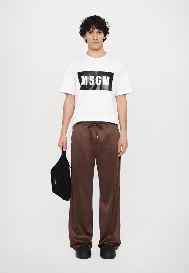 Hvid t-shirt med sort 'MSG'-logo, brune satin drawstring bukser, sort taske og sort fodtøj, stående mod en ensfarvet baggrund.