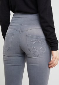 Pantalones vaqueros de mezclilla gris de cintura alta, con dos bolsillos traseros adornados con un patrón de costura decorativa. Textura suave y forma ajustada.