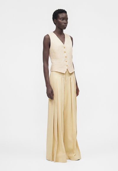 Γυναίκα φορούν μια μπεζ αμάνικη γιλέκο με κουμπιά και κρεμ πλισέ παντελόνια με ψηλή μέση και φαρδιά μπατζάκια, στέκεται μπροστά σε ένα μονόχρωμο φόντο.