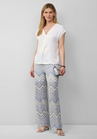 Haut blanc à manches courtes avec un décolleté en V. Pantalon à jambe large avec un motif en zigzag bleu, jaune et blanc. Sac à main argenté et sandales.