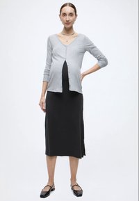 Γκρι ζακέτα πάνω από μαύρο φόρεμα εγκυμοσύνης, και τα δύο στενά. Το φόρεμα είναι μέχρι το γόνατο με πλευρικές σκισμές. Μαύρα επίπεδα παπούτσια ολοκληρώνουν την εμφάνιση.