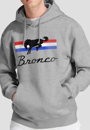Mann trägt hellgrauen Kapuzenpullover mit schwarzer Bucking-Horse-Grafik über roten, weißen und blauen Streifen und dem Text "Bronco" auf der Vorderseite.