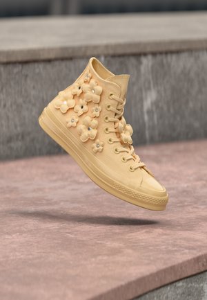 Baskets montantes beige ornées de fleurs en tissu 3D avec de petits strass, flottant au-dessus d'une surface en pierre.