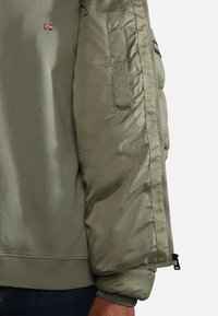 Giacca verde oliva chiaro realizzata in un tessuto misto con una finitura satinata liscia, con polsini a costine e una toppa sul petto con un emblema di bandiera.