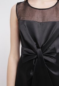 Robe noire avec un haut transparent et un corsage en satin. Présente un nœud proéminent à la taille. Texture lisse avec une silhouette ajustée.
