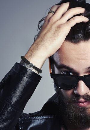 Veste en cuir noir, bracelet en perles noires avec un logo, lunettes de soleil avec des accents orange, et une barbe foncée de longueur moyenne. Main sur la tête, arrière-plan texturé.