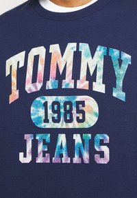 Mörkblå t-shirt i bomull med rund halsringning. Har ett färgglatt tie-dye-mönster på "TOMMY" och "JEANS" med "1985" i en cirkel.