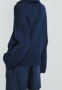 Marineblauer Strickpullover mit weitem Kragen, geripptem Saum und fallenden Schultern, kombiniert mit dunklen Jeans-Shorts mit seitlichen Taschen.