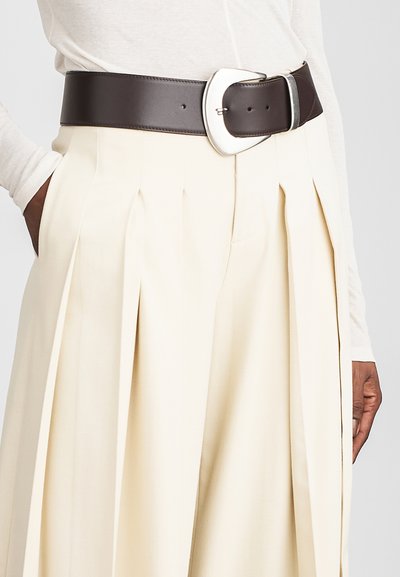 Bruine leren riem met een grote, zilveren ovale gesp, gedragen over crèmekleurige geplooide broeken, die de glatte textuur en brede ontwerp benadrukt.