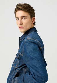 Sininen farkkutakki, jossa on jämäkkä muotoilu. Takissa on viistot kaulus, etutaskut ja näkyvät ompeluyksityiskohdat neutraalilla taustalla.