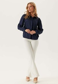 Blusa navy con maniche a palloncino, chiusura con bottoni e texture liscia, abbinata a pantaloni bianchi a zampa d'elefante e tacchi chiari. Design semplice.