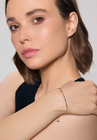 Złota bransoletka o skręconym wzorze z małymi koralikami, noszona na nagim nadgarstku na jednolitym tle.