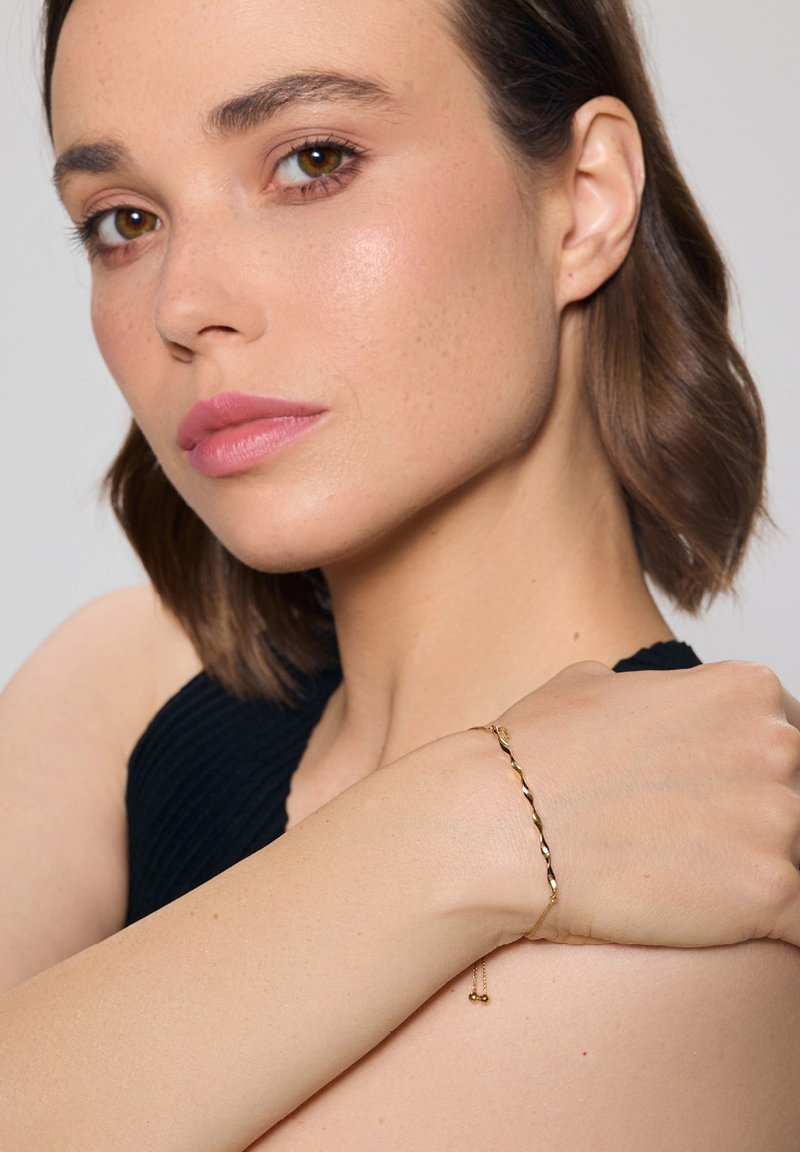 Złota bransoletka o skręconym wzorze z małymi koralikami, noszona na nagim nadgarstku na jednolitym tle.