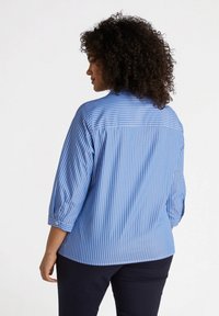 Blusa a rayas azul y blanca con mangas de tres cuartos, detalle de puños con botón y un ajuste relajado. Tela suave con una textura ligeramente crujiente.