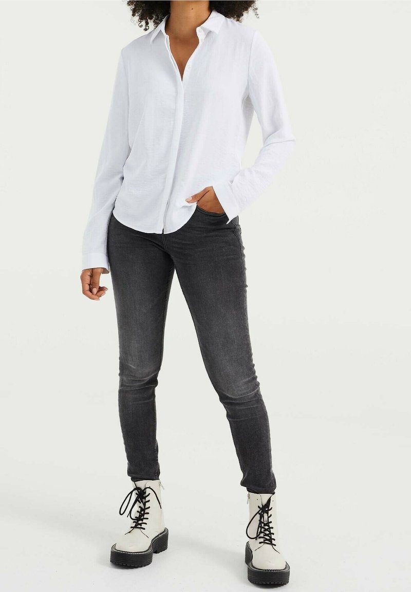 Bílá dlouhá košile s knoflíky, doplněná o těsné tmavě šedé džíny a bílé šněrovací kotníkové boty. Hladká látka a kontrastní textury.