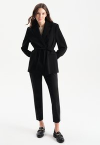Czarna, dopasowana marynarka z wiązanym paskiem w talii, klapami i dwiema przednimi kieszeniami, zestawiona z dopasowanymi spodniami oraz czarnymi loafersami.
