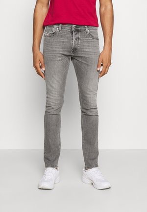 Mann trägt eng anliegende, ausgebleichte graue Jeans, rotes Hemd und weiße Turnschuhe, steht vor einem schlichten weißen Hintergrund.