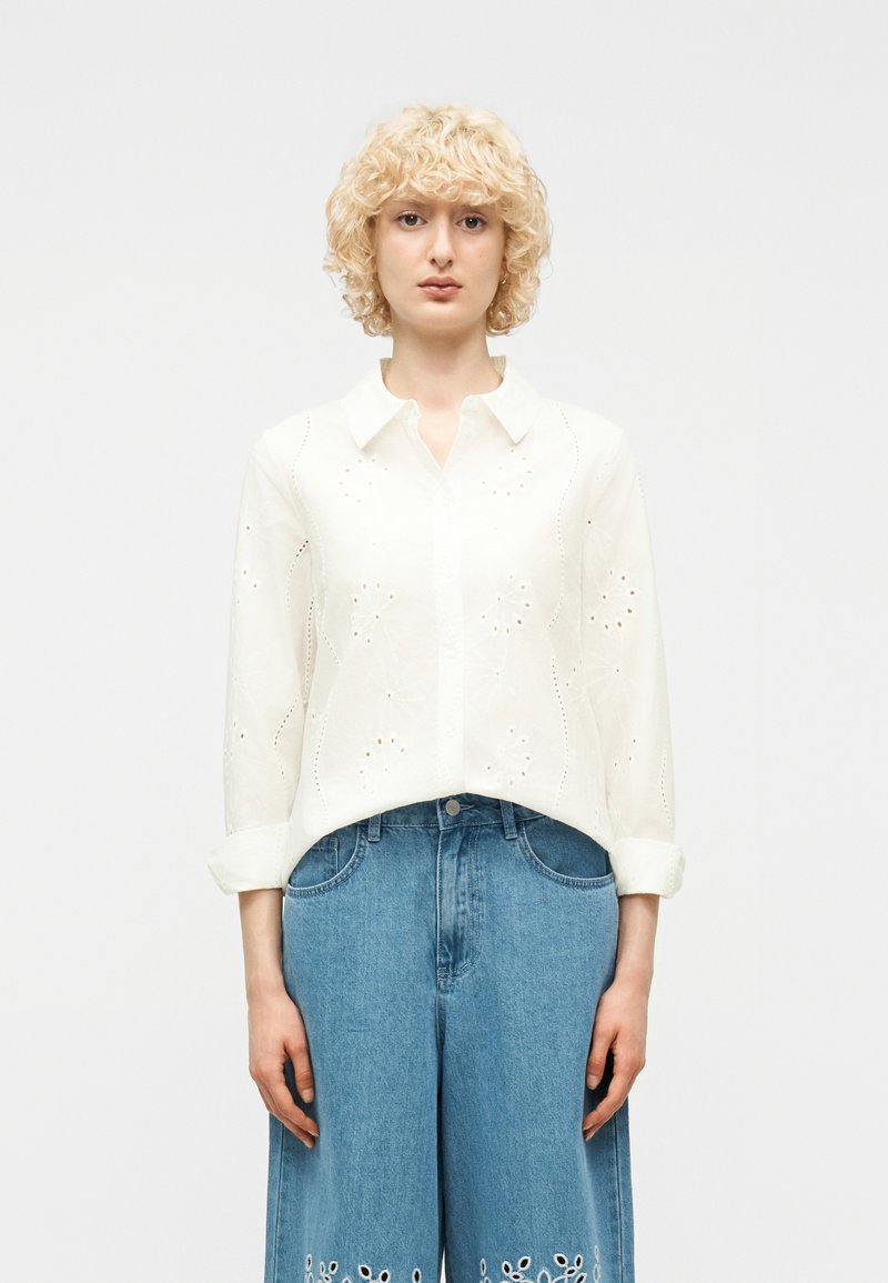 Γυναίκα με σγουρά ξανθά μαλλιά που φοράει λευκό πουκάμισο με κουμπιά και μάτια και ψηλόμεσα μπλε τζιν με λουλουδάτες κοψίματα.