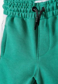 Zaļi kokvilnas joggeri ar sašaurinātu jostasvietu, melnu auklu, sānu kabatām un teksturētu audumu. Akcenti ietver baltus cilpas uz sāniem.