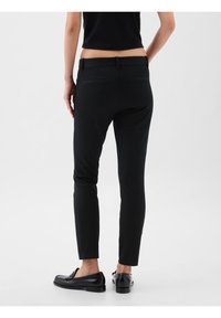 Personne debout, portant un pantalon noir slim-fit et des mocassins noirs, vue de dos sur un fond clair uni.