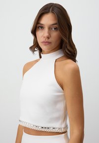 Haut court sans manches blanc avec un col montant, ornée de perles et de sequins le long de l'ourlet. Texture lisse.