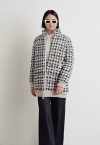 Grau-Hahnentritt-Musterjacke mit hohem Kragen, Frontreißverschluss und gerippten Bündchen, kombiniert mit schwarzen weit geschnittenen Hosen und weißen Brillen.