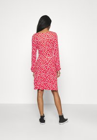 Czerwona sukienka z długimi rękawami, zdobiona białym kwiatowym wzorem, dopasowana w talii, z rozkloszowanym dołem, noszona z czarnymi sneakersami za kostkę.