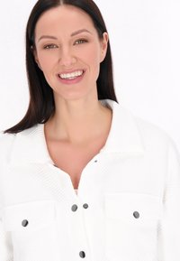 Giacca bianca trapuntata con un motivo testurizzato, dotata di colletto, doppie tasche sul petto e bottoni a pressione neri. Donna sorridente, sfondo chiaro.