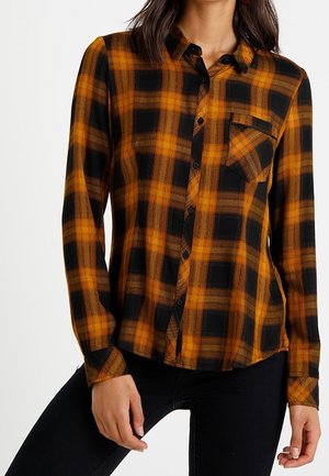 Mujer con una camisa de cuadros naranja y negra de manga larga con bolsillo en el pecho, combinada con pantalones negros.