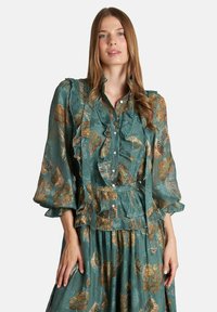 Vestido floral verde con volantes, detalles de botones y mangas abullonadas. La tela tiene una textura transparente con acentos florales metálicos.