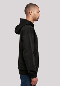 Felpa nera con cappuccio caratterizzata da una vestibilità rilassata, polsini a coste e una texture liscia. Presentata in vista laterale su uno sfondo neutro.