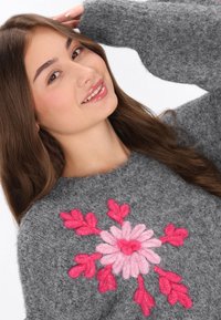 Grått luddigt tröja med en stor blommönster med rosa och ljusrosa petaler, med texturerade garn accenter och en bred halsringning.